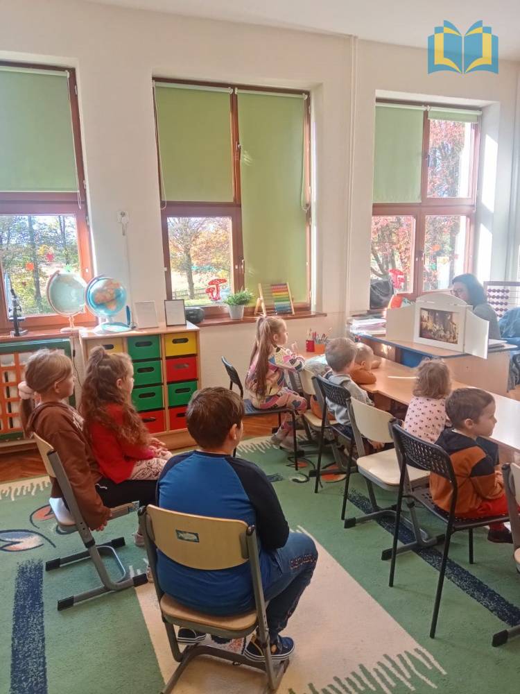Zdjęcie: Klasa szkolna. Dzieci siedzą na krzesłach, słuchają bajki czytanej przez bibliotekarza, zaprezentowanej za pomocą teatrzyku kamischibai.